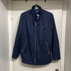 Mörkblå jacka från GANT - Snygg mörkblå jacka från GANT med hög krage och raka linjer. Jackan har två framfickor med dragkedja och diskret GANT-logga på bröstet. Perfekt för dig som gillar stilren och enkel design. Materialet känns slätt och tåligt.