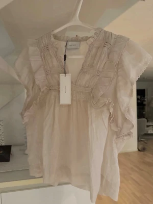 Vit volangblus från Neo Noir - Supersöt vit blus från Neo Noir med volangdetaljer och smockade partier framtill. Blusen har en luftig och romantisk känsla med korta ärmar och rund halsringning. Tillverkad i ett tunt, lätt material som passar perfekt till sommaren.