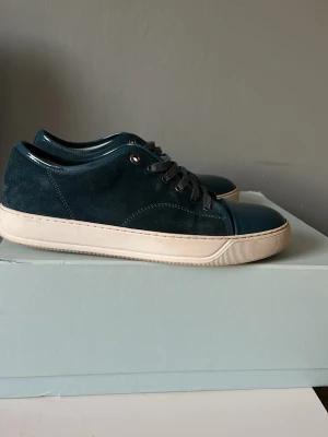 Mörkblå sneakers från Lanvin - Snygga mörkblå sneakers från Lanvin med ovandel i mocka och blank tå. Perfekta för dig som gillar stilrena och exklusiva sneakers. Skorna är inte använda så mycket och har inte någon skada på mockan. Skosnörena skulle behöva bytas för dom är lite smutsiga och jag skickar med ett par helt nya skosnören som är till modellen. Pris går att diskutera!