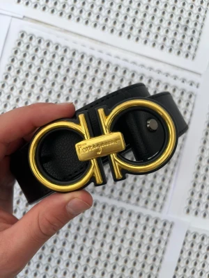 Svart Ferragamo bälte med gulddetalj - Snyggt svart bälte från Salvatore Ferragamo med ikoniskt dubbel-Gancini-spänne i guldmetall. Bältet är gjort i slätt läder och har en exklusiv känsla med Ferragamo-loggan ingraverad på spännet. Perfekt accessoar för att lyfta din outfit.