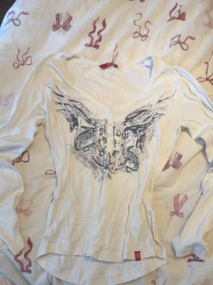 Vit långärmad topp med tryck - Säljer en vit långärmad topp från Replay med ett stort grafiskt tryck i svart och grått framtill. Toppen har en djup v-ringning och figurnära passform. Perfekt för dig som gillar unika detaljer och streetstyle.