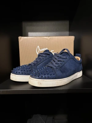 Blå Christian Louboutin sneakers med nitar - Snygga mörkblå sneakers från Christian Louboutin i mocka med ikoniska nitar över hela tån och sidorna. Skorna har klassisk vit sula och den välkända röda undersidan. Perfekt för dig som vill sticka ut med en edgy och lyxig stil.