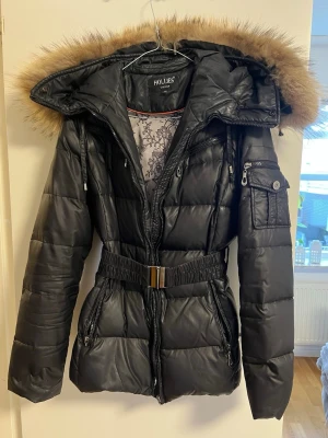 Svart dunjacka med äkta päls från Hollies - Snygg svart dunjacka från Hollies med avtagbar huva och äkta päls. Jackan har markerad midja med bälte, flera fickor med dragkedja och quiltad design. Perfekt för kalla vinterdagar och ger en riktigt trendig look.