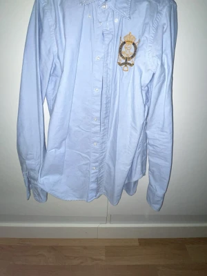 Ljusblå skjorta från Ralph Lauren - Snygg ljusblå skjorta från Ralph Lauren med klassisk krage och guldbroderad logga på bröstet. Skjortan har knappar framtill och är långärmad. Perfekt för dig som gillar stilrena och tidlösa plagg.