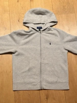 Grå hoodie från Polo Ralph Lauren - Klassisk grå hoodie från Polo Ralph Lauren med dragkedja framtill och huva. Broderad marinblå logga på bröstet och två fickor fram. Tillverkad i mjuk bomull för en skön och avslappnad stil.