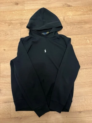 Svart hoodie från Polo Ralph Lauren - Klassisk svart hoodie från Polo Ralph Lauren med huva och den ikoniska vita logobroderingen på bröstet. Tillverkad i mjukt bomullsmaterial och har ribbade muddar vid ärmslut och nederkant. Perfekt för en avslappnad och stilren look.