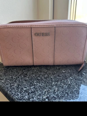 Rosa plånbok från Guess med dragkedja - Snygg rosa plånbok från Guess med silverfärgad logga framtill och präglat mönster. Plånboken har dragkedja runtom, flera kortfack och ett myntfack med dragkedja inuti. Tillverkad i konstläder med stilren design.