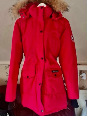 Röd parkas från Canada Goose - Möjlighet att buda eller pruta priset . Snygg röd parkas från Canada Goose med avtagbar huva och äkta pälsdetalj. Jackan har stora fickor framtill, logotyp på ärmen och stängs med dragkedja och knappar. Perfekt för kalla dagar och riktigt varm. Klassisk modell med längre passform. 