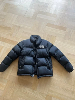 Svart dunjacka från The North Face - Säljer en svart dunjacka från The North Face med hög krage och dragkedja framtill. Jackan har två snedställda fickor med dragkedja och en broderad logga på bröstet. Klassisk boxig passform och tjock vaddering för extra värme. Obs barnstorlek 