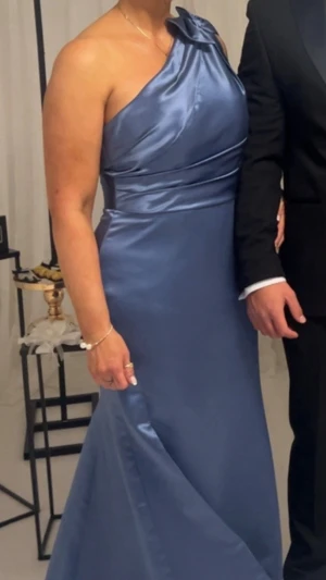 Lång blå balklänning i satin - Elegant lång klänning i skimrande blå satin med figurnära passform och utsvängd kjol. Perfekt för bal eller festliga tillfällen. Klänningen har en enkel och stilren design utan synliga detaljer eller mönster.