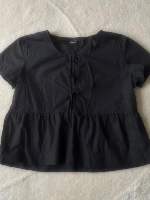 Svart blus med snörning - Säljer en svart blus från Gina Tricot med korta ärmar och rund halsringning. Toppen har ett sött knyt framtill och en lätt volang nedtill som ger en avslappnad look. Perfekt att styla med jeans eller kjol Storlek M men passar bättre på S!💕