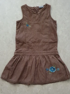 Klänning - Söt brun ärmlös klänning i manchestertyg med blå blombroderier på bröstet och kjolen. Klänningen har en markerad midja och lätt utställd kjol. Perfekt för dig som gillar retrovibbar och detaljer.