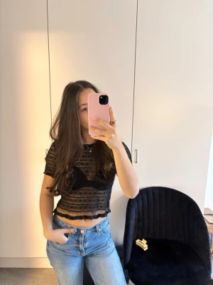 Svart spetsig croppad topp - Säljer en svart, croppad topp i spets med korta ärmar och vågig volangkant nedtill. Toppen är genomskinlig och har ett snyggt mönster i spetsen. Perfekt att styla över ett linne eller bh för en trendig look.