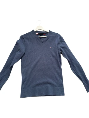 Mörkblå V-ringad tröja Tommy Hilfiger - Klassisk mörkblå stickad tröja från Tommy Hilfiger med V-ringning och liten logga på bröstet. Tröjan har långa ärmar och ribbade muddar vid ärmslut och nederkant. Perfekt för en stilren och enkel look. Pris kan diskuteras, passar för den som är runt 162/172cm lång. Hör av er vid fler frågor.