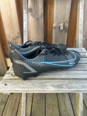 Nike Mercurial svarta fotbollsskor - Säljer ett par svarta Nike Mercurial fotbollsskor med blå detaljer och vita grafiska linjer. Skorna har snörning, låg profil och dobbar för spel på gräs. Tillverkade i syntetmaterial för bra grepp och kontroll på planen. Perfekta för dig som vill ha stilrena och snabba skor.