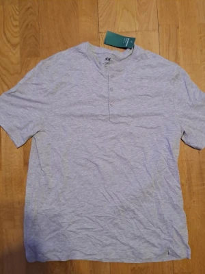 Ljusgrå henley t-shirt från H&M - Ljusgrå kortärmad henley t-shirt från H&M med tre knappar vid halsen. Tillverkad i mjukt bomullsmaterial och har en avslappnad passform. Perfekt för en enkel och stilren look.
