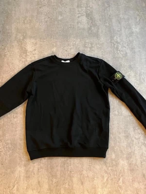 Svart sweatshirt från Stone Island - Säljer en svart sweatshirt från Stone Island med den ikoniska loggan på vänster ärm. Tröjan har rund halsringning, ribbade muddar och är tillverkad i mjukt bomullsmaterial. Perfekt för en clean och stilren look.
