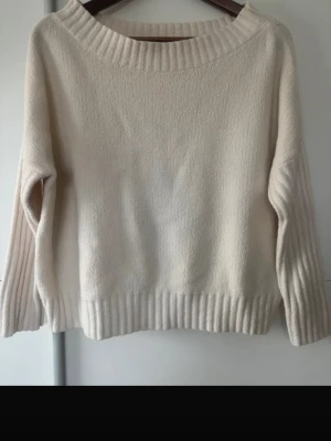 Krämvit stickad tröja med ribbade detaljer - Mysig krämvit stickad tröja med bred ribbad kant vid hals, ärmslut och nederkant. Tröjan har en avslappnad passform och långa ärmar. Perfekt för lager på lager och enkel att matcha med olika outfits.