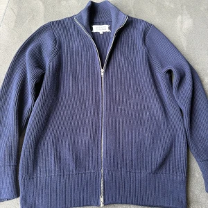 Marinblå stickad ziptröja Maison Margiela - Snygg marinblå stickad tröja från Maison Margiela med hel dragkedja framtill och hög krage. Tröjan har en ribbstickad struktur och raka ärmar. Perfekt för lager på lager och stilrena outfits.