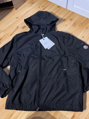 Svart vindjacka från Moncler - Snygg svart vindjacka från Moncler med huva och dragkedja framtill. Jackan har två fickor med dragkedja, elastiska muddar och en Moncler-logga på ärmen. Tillverkad i lätt och slitstarkt material som står emot vind. Perfekt för dig som vill ha en stilren och funktionell jacka.