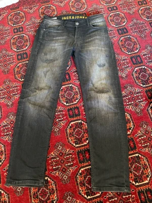 Svarta slitna jeans från Jack & Jones - Säljer ett par svarta jeans från Jack & Jones med slitningar och tvättade detaljer. Jeansen har smal passform, klassisk femficksdesign och knappgylf. Materialet är stretchigt denim som ger en bekväm känsla och snygg look.