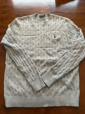 Beige kabelstickad tröja Polo Ralph Lauren - Kabelstickad grå tröja från Polo Ralph Lauren med klassisk svart logga på bröstet. Tröjan har rund halsringning, långa ärmar och ribbade muddar vid ärmslut och nederkant. Perfekt för lager på lager och stilren look.