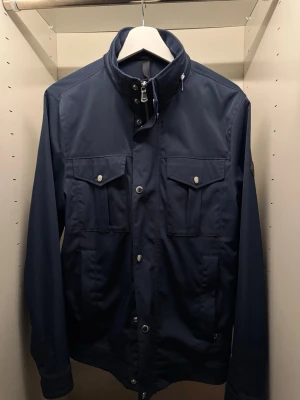 J.Lindeberg Jacka Bailey - Säljer en riktigt fet J.Lindeberg jacka i en fin blå färg, tunn men varm med mycket fickor och inbyggd luva i nack kragen, perfekt nu till vår/sommar! Skicket är 9-10 och den är köpt på Wallenberg för 3500 kr. Storleken är S men passar som M även. Kom PM för mer information/frågor! 🗣️ KAN GÅ NER I PRIS VID SNABB AFFÄR! 🤝🏻