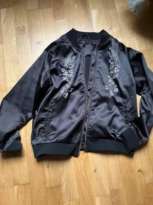 Svart satin bomberjacka med brodyr - Snygg svart bomberjacka i glansigt satinmaterial med broderade detaljer framtill i form av fåglar och blommor. Jackan har ribbade muddar vid ärmslut och nederkant samt dragkedja framtill. Perfekt för dig som vill ha en unik och trendig look.