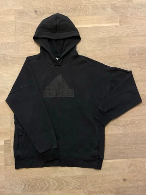 Svart hoodie från Adidas med logga - Svart hoodie från Adidas med stor, ton-i-ton logga på bröstet. Klassisk modell med huva och ribbade muddar vid ärmslut och nederkant. Perfekt för en avslappnad och sportig stil. Mjuk bomullsblandning för extra komfort.