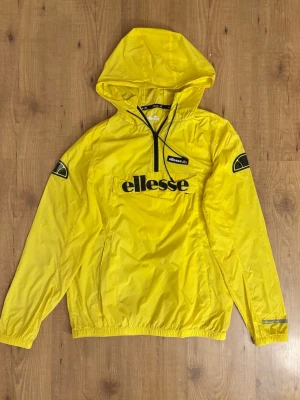 Gul vindjacka från Ellesse med huva - Snygg gul vindjacka från Ellesse med svart logga på bröstet och ärmarna. Jackan har huva, halv dragkedja framtill och elastiska muddar. Tillverkad i lätt och tåligt material som passar perfekt för blåsiga dagar.