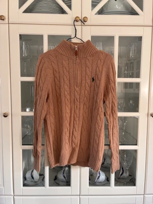 Beige kabelstickad tröja Polo Ralph Lauren - Säljer en beige kabelstickad tröja från Polo Ralph Lauren med halvzip och hög krage. Tröjan har ett broderat mörkblått logotyp på bröstet och ribbstickade muddar. Perfekt för dig som gillar klassisk och stilren design.
