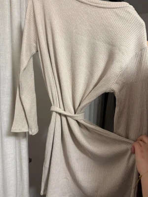 Beige ribbstickad klänning med knytband - Säljer en beige ribbstickad klänning med långa ärmar och ett matchande knytband i midjan. Klänningen har en mjuk och stretchig känsla, perfekt för lager på lager. Enkel och stilren design utan synligt märke.