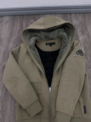 Olivgrön hoodie moose knuckles med foder - Säljer en olivgrön hoodie från moose knuckles med mjukt teddyfoder i huvan och insidan. Hoodien har dragkedja, två fickor och en patch på ärmen. Perfekt för dig som vill ha något både snyggt och bekvämt. Ribbade muddar och detaljer i svart.