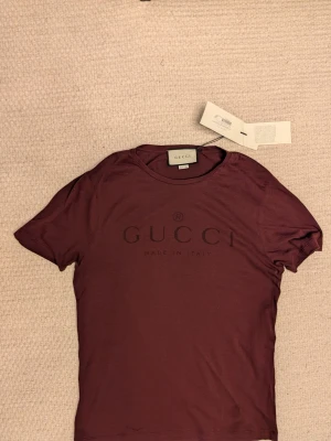 Oanvänd Gucci t-shirt - Snygg vinröd t-shirt från Gucci med diskret logga och texten 'Made in Italy' på bröstet. Klassisk rund halsringning och korta ärmar. Tillverkad i mjuk bomull som känns skön mot huden. Perfekt för dig som vill ha en stilren och exklusiv look.