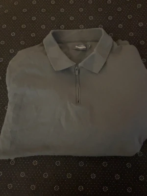Grå polotopp med dragkedja Dressman - Snygg grå polotopp från Dressman med klassisk krage och halv dragkedja framtill. Tillverkad i mjukt material som känns skönt mot huden. Perfekt till jeans eller kjol för en clean och enkel look.