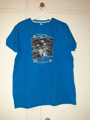 Blå t-shirt med grafiskt tryck Trespass - Blå t-shirt från Trespass med ett stort grafiskt tryck på bröstet i svart och vitt med texten 'DOWNHILL' och topografiska linjer. Rund halsringning och klassisk passform. Perfekt för dig som gillar streetwear och coola prints.