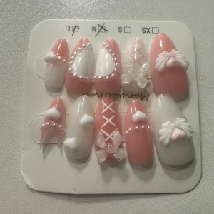 Söta rosa och vita press-on naglar den sista längst ner är halvt sönder  - Supergulliga press-on naglar i rosa och vitt med pärldetaljer, hjärtan och 3D-rosetter. Några naglar har korsade band och transparenta dekorationer för extra wow-effekt. Perfekt för dig som vill ha en unik och söt nagelstil.