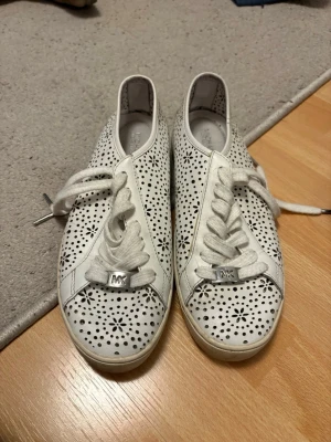 Vita sneakers från Michael Kors - Snygga vita sneakers från Michael Kors med blommigt perforerat mönster över hela skon. Låga i modellen med vita snören och metallisk MK-detalj framtill. Tillverkade i läder med gummisula och diskret logga på sidan av sulan. Perfekta för dig som gillar stilrena men ändå unika skor.