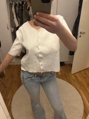 Vit kortärmad cardigan med knappar - Säljer en vit, kortärmad cardigan med v-ringning och knappar framtill. Toppen är croppad och har en rak passform. Materialet ser ut att vara mjukt och lätt ribbat, perfekt för en avslappnad och trendig look.