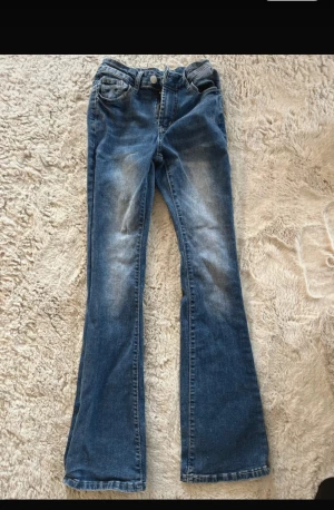 Blå bootcut jeans med slitningar - Ett par blå bootcut jeans med snygga slitningar framtill och bakfickor. Jeansen har klassisk femficksmodell, normal midja och är tillverkade i denim. Perfekta för dig som gillar en retroinspirerad look med utsvängda ben. Mycket bra skick.