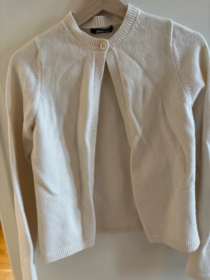 Krämvit kofta från Gina Tricot - Stilren krämvit kofta från Gina Tricot med rund halsringning och ribbade detaljer. Koftan har en knapp framtill vid halsen och är tillverkad i mjukt stickat material. Perfekt att slänga över en topp för en enkel och snygg look.