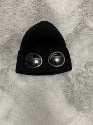  C.P. Company mössa - Cool svart mössa från C.P. Company med inbyggda glasögon i uppviket. Stickad design och rund form, perfekt för att ge en unik och trendig look. Glasögonen har märkets logga på ena linsen och ger mössan en futuristisk vibe.