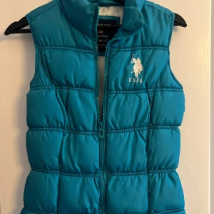 Turkos dunväst från U.S. Polo Assn. - Snygg turkos dunväst från U.S. Polo Assn. med hög krage och dragkedja framtill. Västen har broderad logga på bröstet och är tillverkad i 100% polyester. Perfekt för lager på lager och ger en sportig vibe till din outfit.