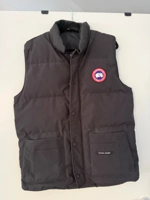 Svart dunväst från Canada Goose - Snygg svart dunväst från Canada Goose med klassisk logga på bröstet. Västen har hög krage, två stora fickor framtill och stängs med både dragkedja och tryckknappar. Perfekt för lager på lager och håller dig varm under kyliga dagar.
