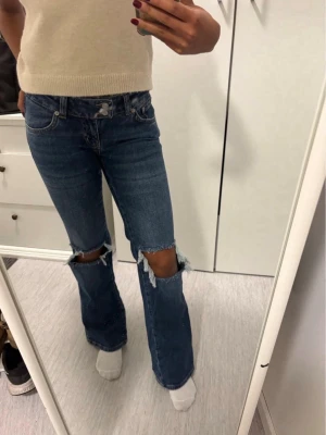 Blå bootcut jeans med stora hål - Säljer ett par blå bootcut jeans med stora slitna hål över knäna. Jeansen har klassisk femficksdesign och är tillverkade i denim. Perfekta för dig som gillar en trendig och avslappnad stil.💕
