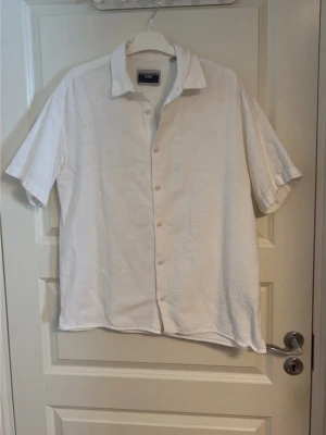 Vit kortärmad skjorta från Jack & Jones - Säljer en stilren vit skjorta från Jack & Jones med korta ärmar och klassisk krage. Skjortan har knappar framtill och är gjord i ett lätt bomullstyg, perfekt för varma dagar. Enkel och clean look som passar till det mesta.