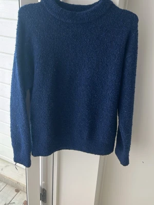 Mörkblå stickad tröja - Mysig mörkblå stickad tröja med rak passform och rund halsringning. Tröjan har långa ärmar och är tillverkad i ett mjukt, strukturerat material som ger en avslappnad look. Perfekt för kyliga dagar och enkel att matcha med jeans eller kjol.