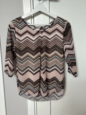 Mönstrad topp med zigzag och trekvartsärm - Säljer en snygg topp med zigzag-mönster i beige, grön, brun, svart och vitt. Blusen har trekvartsärm och rund halsringning med liten knytning bak. Materialet känns lätt och luftigt, perfekt för dig som gillar grafiska mönster.
