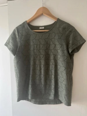 Grön topp från Jacqueline de Yong - Snygg grön topp från Jacqueline de Yong med korta ärmar och ett coolt hålmönster i geometrisk stil. Toppen har rund halsringning och är gjord i mjuk bomull, perfekt för en avslappnad look.