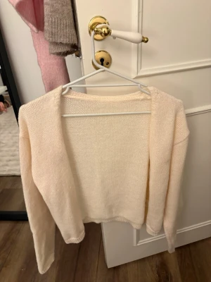Ljusbeige cropped stickad cardigan - Säljer en ljusbeige cropped stickad cardigan med lång ärm och öppen framtill. Perfekt att slänga över en topp för en chill och stilren look. Mjuk och tunn stickning som ger en avslappnad vibe.
